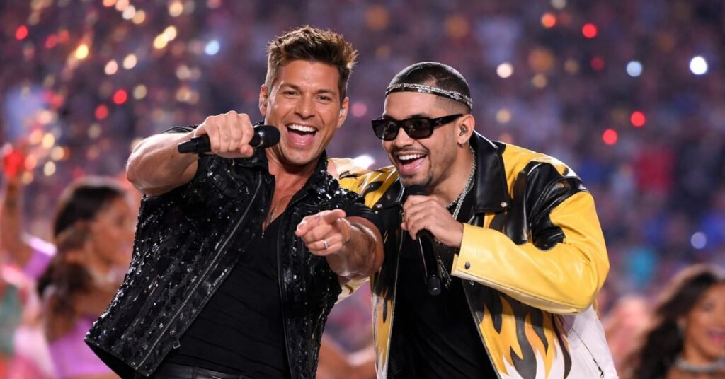 Ricky Martin and Bad Bunny perform together on a brightly lit Super Bowl halftime stage, smiling and singing into microphones as colorful lights and dancers blur in the background.