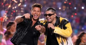 Ricky Martin and Bad Bunny perform together on a brightly lit Super Bowl halftime stage, smiling and singing into microphones as colorful lights and dancers blur in the background.